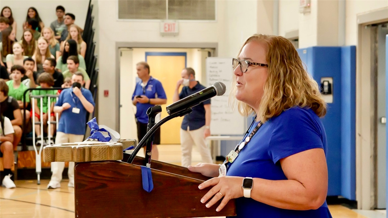   Kelley St. Coeur wears a blue shirt as she stands at a podium in the AHS gym 