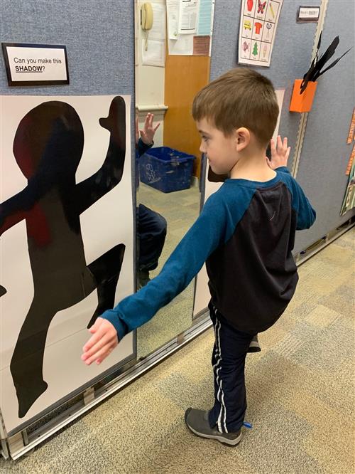 Young Boy Raising LEg and bending arms so mirror his body in the same shape as a wall shadow 